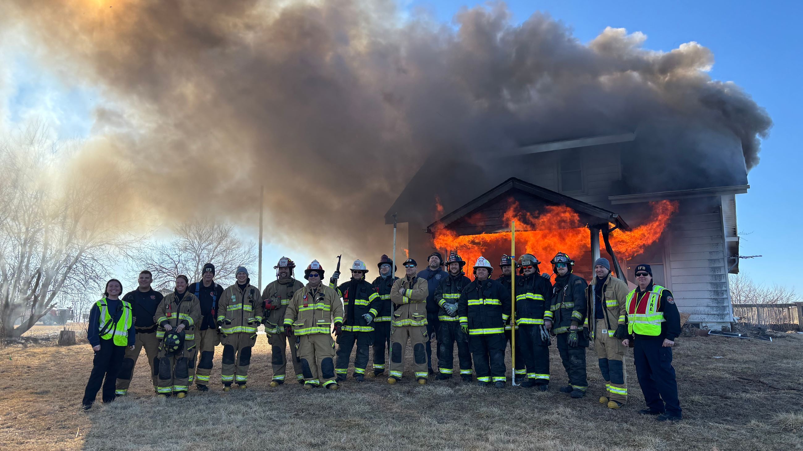A group of firefighters stand in front of a burning structure during a training exercise.