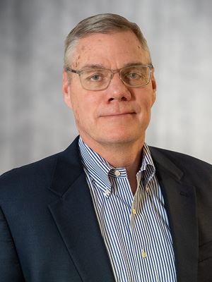 A man in glasses and a blue suit smiles for a photo in front of a grey backdrop.