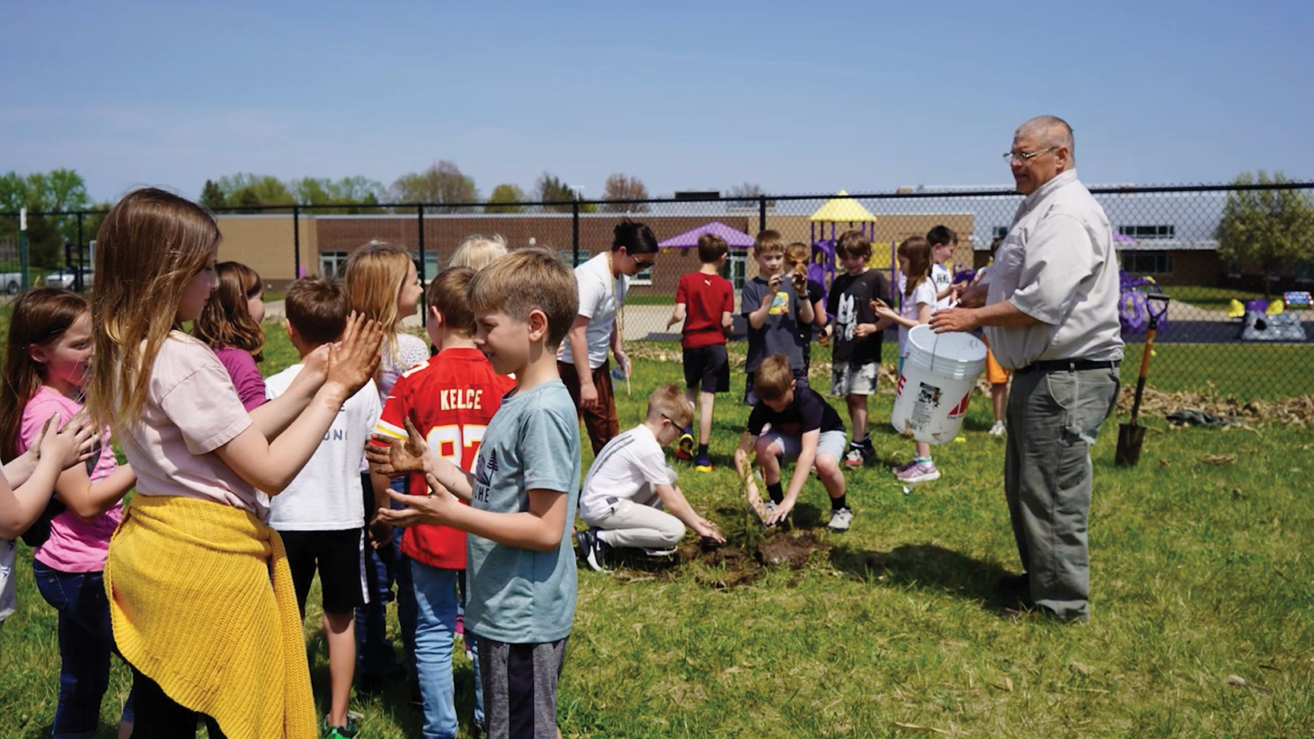 A group of students and a man in glasses plant trees outside a school playground.