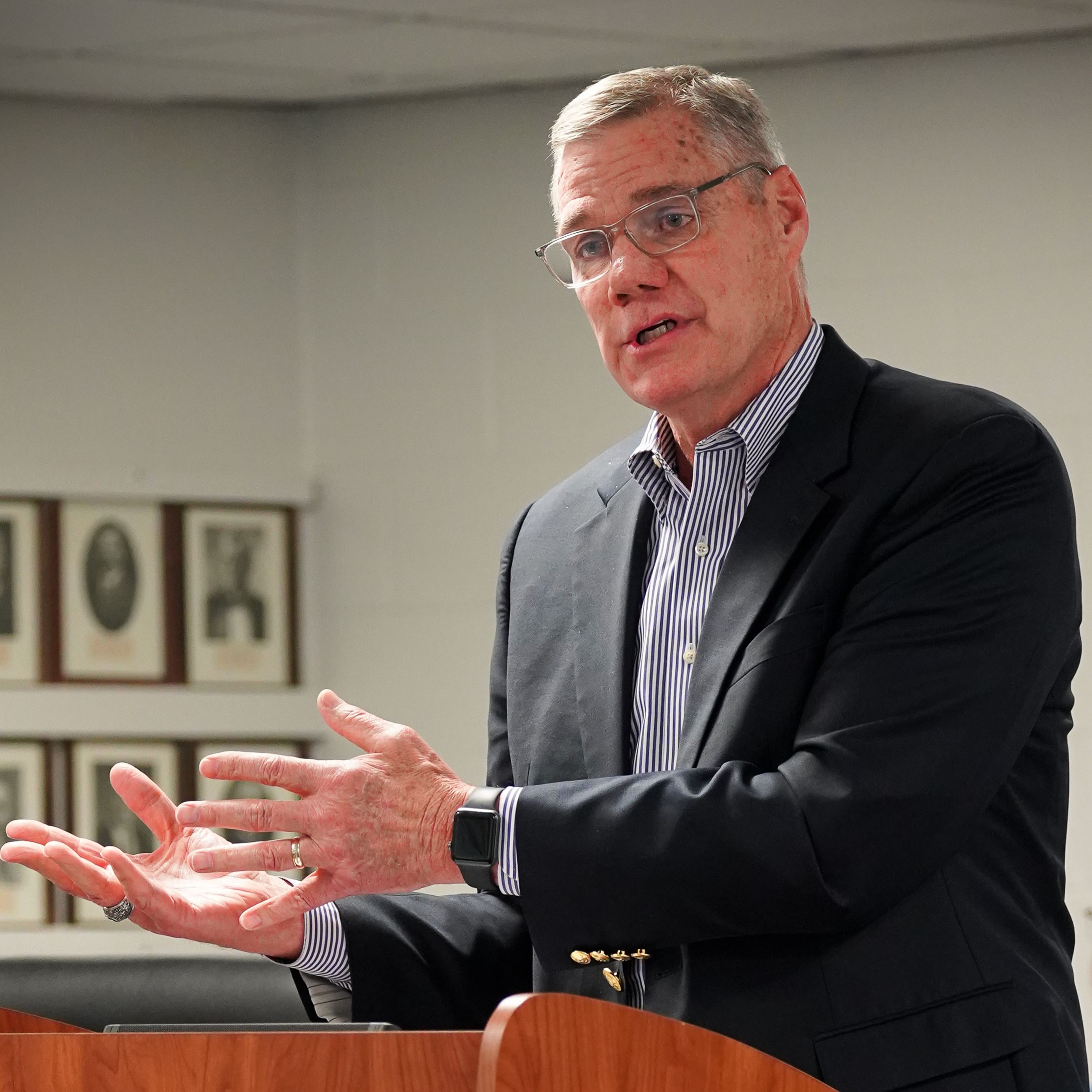 A man wearing a suit and glasses speaks to people while standing at a podium.