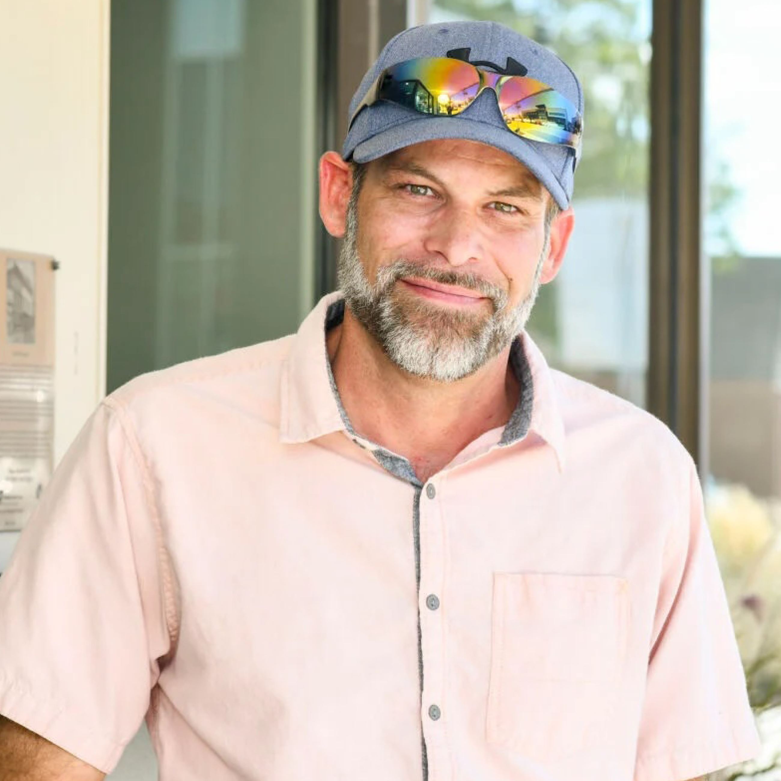 A man wearing a hat and a cream-colored shirt smiles for a portrait photo in front of a building.