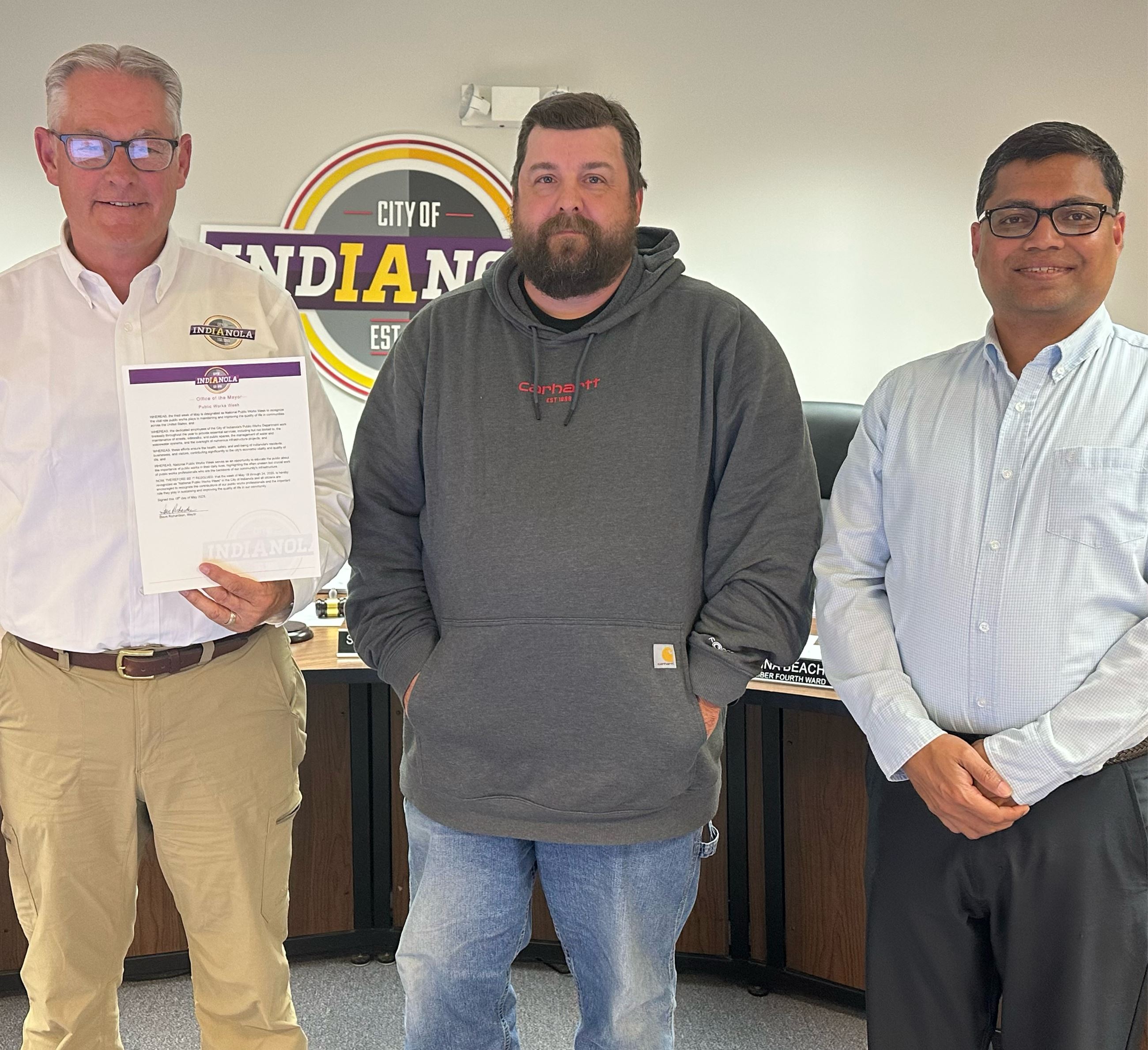 Three men pose for a photo holding a proclamation declaring May 18-24 as Public Works Week in Indian
