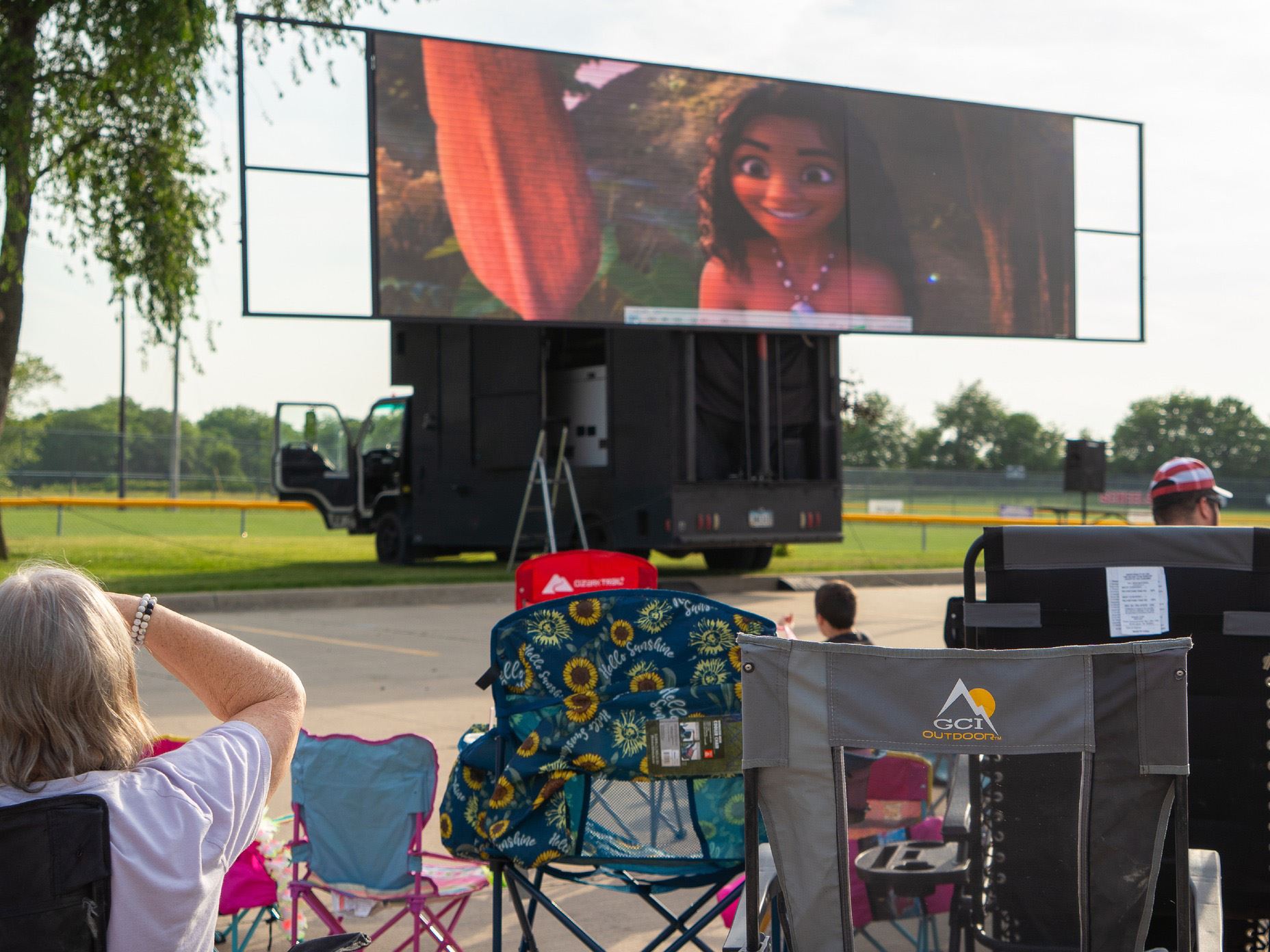 People sitting at a ballpark in lawn chairs watch a movie on a giant screen.
